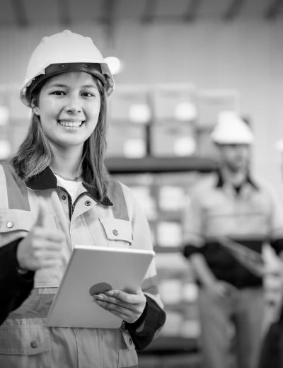 Warehouse worker young woman hold tablet with show thumbs up and look at camera and smiling with her coworkers discuss in the background.
